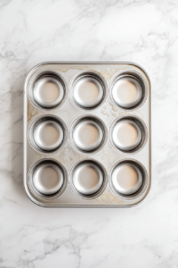 This image shows a nonstick 12-cup metal muffin tin placed on a white marble countertop, each cup evenly coated with nonstick spray, ready for the hash brown mixture.