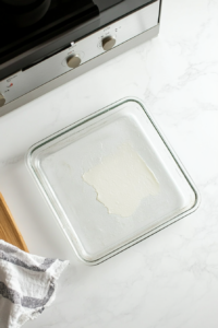 This image shows a top-down view of a lightly greased square glass baking pan on a white marble cooktop with an oven preheating in the background at 350°F.
