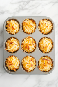 This image shows a metal muffin tin over a white marble countertop, with each cup filled and pressed with the seasoned shredded potato mixture to form uniform basket shapes.
