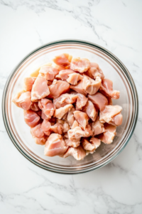 This image shows raw, skinless cut-up chicken pieces placed inside a large clear glass bowl, set on a clean white marble countertop, with no other items or ingredients in the background.