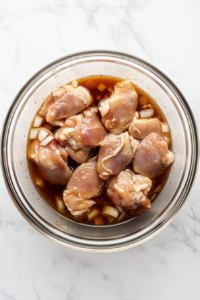 This image shows raw chicken thighs soaking in a dark soy and coconut marinade in a glass bowl placed on a clean white marble countertop.