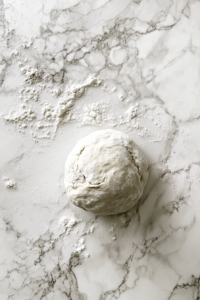 This image shows a dough ball resting on a lightly floured marble surface, covered with a glass bowl.