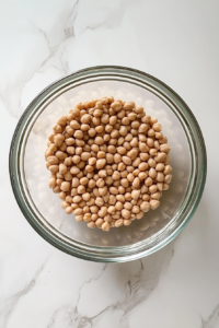 This image shows a top-down view of a clear glass bowl on a white marble countertop, filled with thoroughly rinsed, drained, and dried chickpeas and navy beans, ready for the salad.