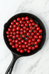 This image shows a black cast iron skillet on a clean white marble countertop, filled with cherry tomatoes drizzled with olive oil and sprinkled with thyme leaves, roasting gently.