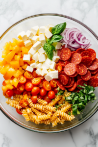 This image shows Italian dressing drizzled over rotini pasta and mixed vegetables in a glass bowl on a white marble cooktop.