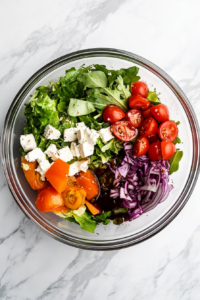 This image shows salad dressing and balsamic vinegar added over layered autumn salad ingredients in a glass bowl on a white marble cooktop.