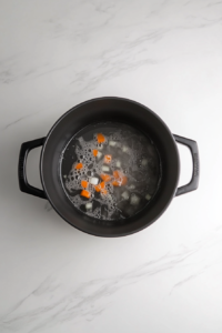 This image shows chopped onion, carrots, and celery sautéing in melted butter inside a large black soup pot on a white marble countertop.