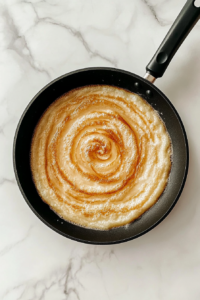 This image shows a black skillet with sautéing onion, garlic, and ginger paste in oil, sitting on a white marble cooktop.