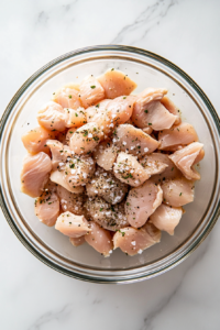This image shows bite-size raw chicken pieces seasoned with salt, black pepper, and oregano in a large clear glass bowl, sitting on a clean white marble cooktop.