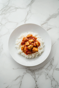This image shows a serving of chicken curry and white rice on a plate placed on a clean white marble cooktop.