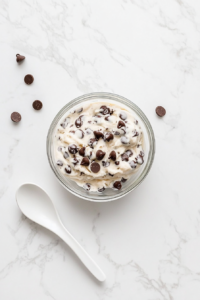 This image shows a glass bowl filled with ready-to-eat cookie dough, dotted with chocolate chips, on a spotless white marble countertop with a white spoon beside it.