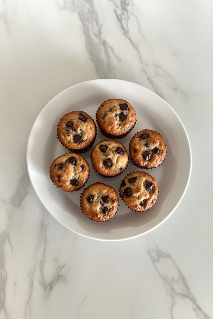 This image shows warm chocolate chip muffins arranged on a white ceramic plate on a white marble countertop, ready to serve.