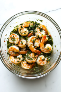 This image shows raw shrimp tossed in chili-lime dressing inside a clear glass mixing bowl on a white marble cooktop. The shrimp glisten with the dressing, and the scene is clean and minimal.