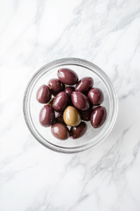 This image shows a top-down view of a small clear glass bowl on a white marble countertop, containing thinly sliced pitted Kalamata olives, ready to be added to the salad.