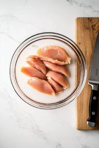 This image shows six thin chicken breast slices in a glass bowl on a white marble surface, beside a knife and cutting board, captured from a top-down angle.
