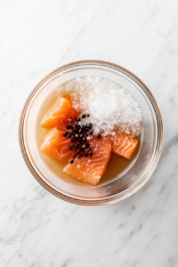 This image shows raw salmon fillets submerged in a brine of water, kosher salt, brown sugar, and black peppercorns inside a clear glass bowl on a white marble countertop.
