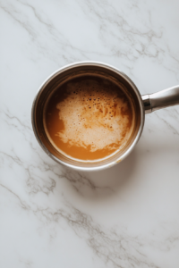 This image shows evaporated milk being added gradually to the bubbling caramel base in a stainless steel saucepan on a white marble cooktop.