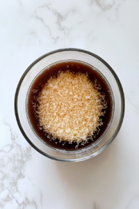 This image shows a glass bowl of Hawaiian marinade with soy sauce, coconut milk, onions, garlic, and other ingredients mixed together on a white marble surface.