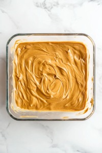 This image shows a top-down view of a square glass baking dish filled with evenly spread peanut butter brownie batter over a white marble cooktop.