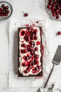This image shows cherry cream being spread evenly over an unrolled chocolate sponge on a parchment sheet, resting on a white marble cooktop.