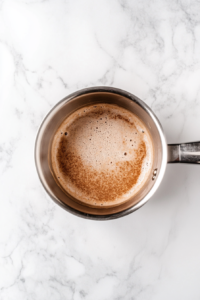 This image shows a steaming mixture of whole milk, heavy cream, powdered sugar, and espresso powder in a stainless steel saucepan on a white marble cooktop, with tiny bubbles forming at the edges.