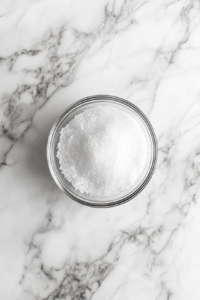 This image shows a top-down view of a small glass bowl filled with granulated sugar on a white marble countertop.