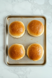 This image shows sandwich buns face-up on a cookie sheet after toasting, placed on a clean white marble cooktop.