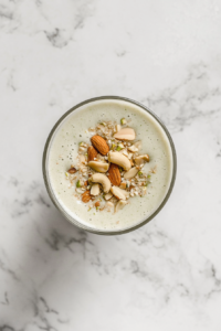 This image shows a tall clear glass filled with avocado banana smoothie, topped with chopped cashews, almonds, and pistachios, sitting on a white marble countertop.