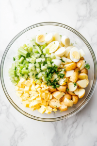 This image shows a clear glass bowl containing gently mixed potato salad with potatoes, celery, onion, eggs, and creamy dressing on a white marble countertop.