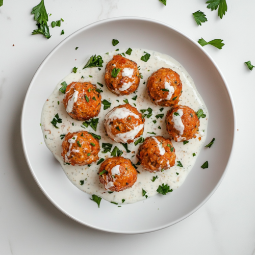 This image shows a plate of golden-brown tuna meatballs arranged neatly with creamy yogurt sauce drizzled on top, garnished with fresh parsley, on a clean white marble countertop.