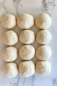 This image shows twelve smooth and oiled dough balls placed on a floured white marble surface.