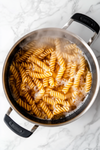 This image shows a black pot on a white marble cooktop with rotini pasta boiling in water, bubbles rising and steam visible, against a clean, simple background.