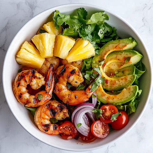 This image shows a vibrant shrimp and pineapple avocado salad arranged in a white ceramic bowl placed on a clean white marble countertop, topped with grilled shrimp, pineapple slices, creamy avocado, halved cherry tomatoes, red onion slices, and romaine lettuce.