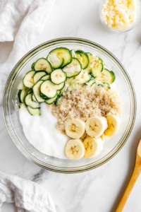 This image shows a glass mixing bowl on a white marble countertop with well-combined ingredients for zucchini banana bread, including beaten eggs, shredded zucchini, mashed bananas, oil, white sugar, brown sugar, and vanilla extract.