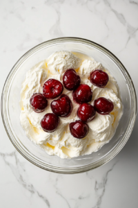 This image shows a glass bowl of whipped cream mixed with icing sugar and chopped cherries on a white marble cooktop for cake filling.