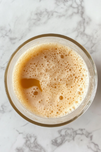 This image shows a well-combined frothy mixture of eggs and sugars in a glass bowl on a clean white marble cooktop.