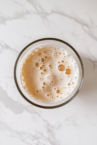 This image shows a glass bowl containing a pale, frothy mixture of egg yolks, sugar, and salt, placed on a clean white marble cooktop.