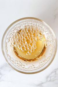 This image shows a large clear glass bowl on a white marble countertop containing warm water, honey, and blooming yeast.