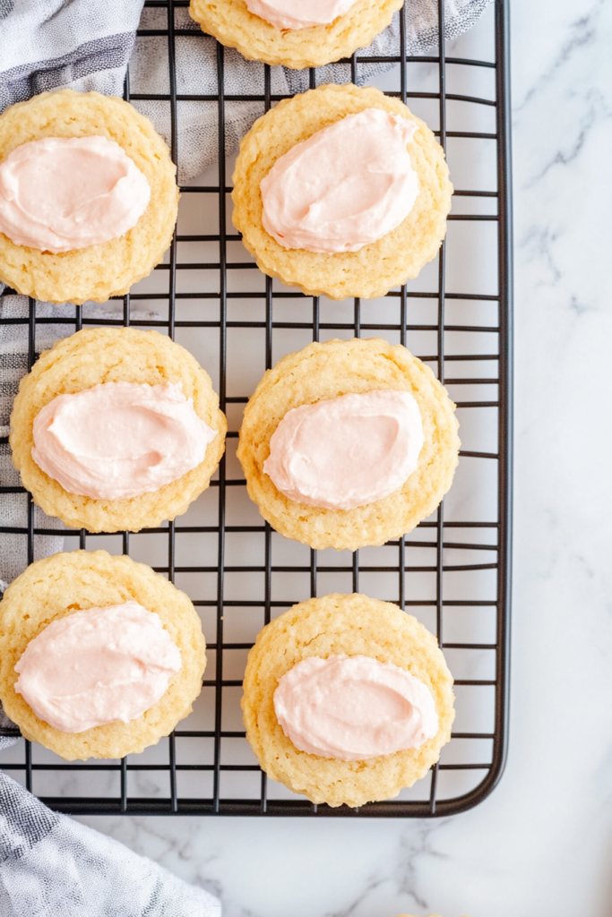 This image shows a top-down view of pale golden yo yo biscuits being assembled on a black wire cooling rack over a clean white marble countertop, with one biscuit spread with pale pink filling and another placed on top to form a sandwich.