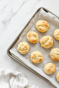 This image shows a top-down view of the silver baking tray with lightly golden yo yo biscuits fresh from the oven, cooling on the parchment paper over a clean white marble countertop.