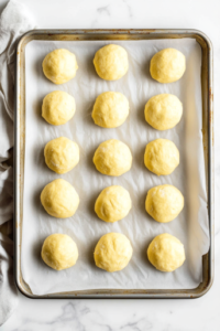 This image shows a top-down view of the silver baking tray lined with parchment paper on a clean white marble countertop, filled with evenly spaced small pale yellow dough balls pressed lightly on top with a fork, ready for baking.