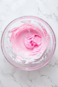 This image shows a top-down view of the same clear glass mixing bowl on a clean white marble countertop, now containing the smooth creamy filling mixed with a drop of pink food coloring, creating a pale pink hue.