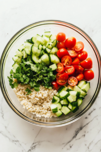 This image shows a clear glass bowl on a white marble cooktop filled with fluffed brown rice and topped with diced zucchini, chopped cucumber, halved cherry tomatoes, sliced green onions, and freshly chopped cilantro.