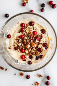 This image shows the zucchini banana bread batter with cranberries and chopped nuts gently folded in, placed in a glass bowl on a white marble countertop.