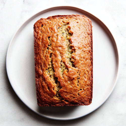 This image shows a close-up shot of a freshly baked zucchini banana bread loaf, slightly sliced to reveal the moist interior with banana and zucchini chunks, placed on a white marble countertop.