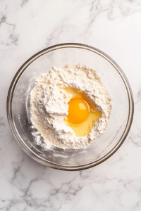 This image shows eggs and vanilla extract being incorporated into a butter-sugar mixture in a clear glass bowl on a white marble surface.