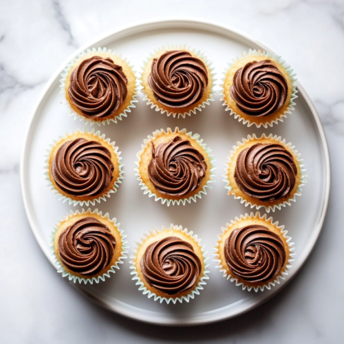 This image shows golden mini vanilla cupcakes with chocolate frosting swirls, neatly arranged on a white plate over a spotless white marble countertop.