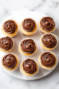 This image shows mini vanilla cupcakes with chocolate frosting beautifully served on a white ceramic plate over a spotless white marble countertop.