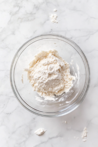 This image shows a clear glass bowl on a white marble countertop with partially mixed milk cookie ingredients and visible added flour, forming a soft, thickening dough.