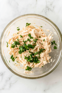 This image shows shredded chicken and parsley being folded into a creamy mixture in a glass bowl on a white marble surface.